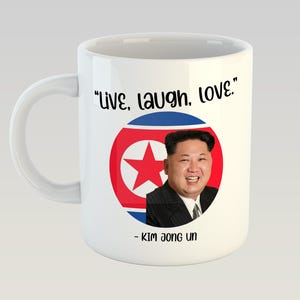 Può includere: Una tazza da caffè in ceramica bianca con un'immagine in bianco e nero di Kim Jong Un davanti a una bandiera nordcoreana. Il testo "live, laugh, love" è stampato in nero sopra l'immagine e "Kim Jong Un" è stampato in nero sotto l'immagine.