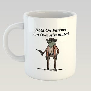 Op de afbeelding: Een witte keramische mok met een zwarte handgreep. De mok heeft een cartoonillustratie van een kikker die een cowboyhoed draagt en een pistool vasthoudt. De tekst "Hold On Partner I'm Overstimulated" staat op de mok gedrukt.