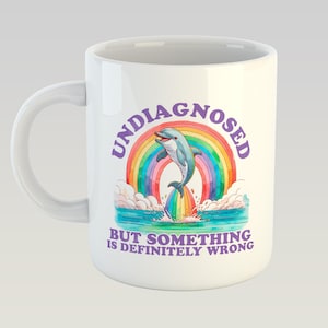 Op de afbeelding: Witte keramische koffiemok met een paarse, regenboogkleurige en blauwe afbeelding van een dolfijn die over een regenboog springt. De tekst "Undiagnosed but something is definitely wrong" staat in paars op de mok gedrukt.