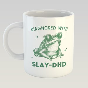 May include: White ceramic mug with a green illustration of a frog wearing sunglasses. The mug has the text "DIAGNOSED WITH" above the frog and "SLAY-DHD" below. The mug has a curved handle.
