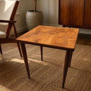 May include: A square side table featuring a light brown burl wood veneer top with a distinct swirling grain pattern. The table is supported by four dark brown, tapered wooden legs that extend outward from the corners of the square tabletop.