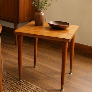 May include: A square wooden side table with a warm brown finish, featuring four tapered legs capped with metallic gold-toned feet. The table surface holds a brown ceramic vase containing green foliage and a small, shallow wooden bowl.