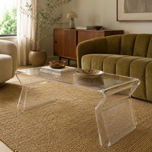 May include: A clear acrylic coffee table with a waterfall edge design. The rectangular table features a smooth, transparent surface and angled side supports. Two stacked books, one titled AALTO, and a small brown bowl sit on the table top.