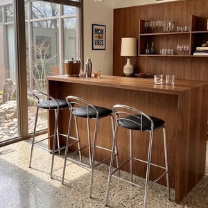 May include: Three bar stools with chrome metal frames and black padded seats positioned in front of a wooden bar counter. The bar features a wooden shelving unit holding glassware, a lamp, a metal ice bucket, and a shaker set on the surface.