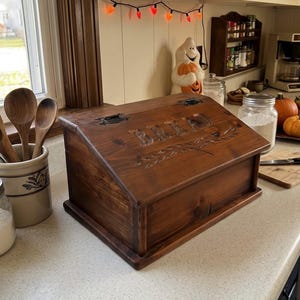 May include: A rectangular wooden bread box with a slanted hinged lid featuring the word BREAD carved into the front panel. The box has a dark brown stained finish, visible wood grain, and black metal hinges on the top lid.