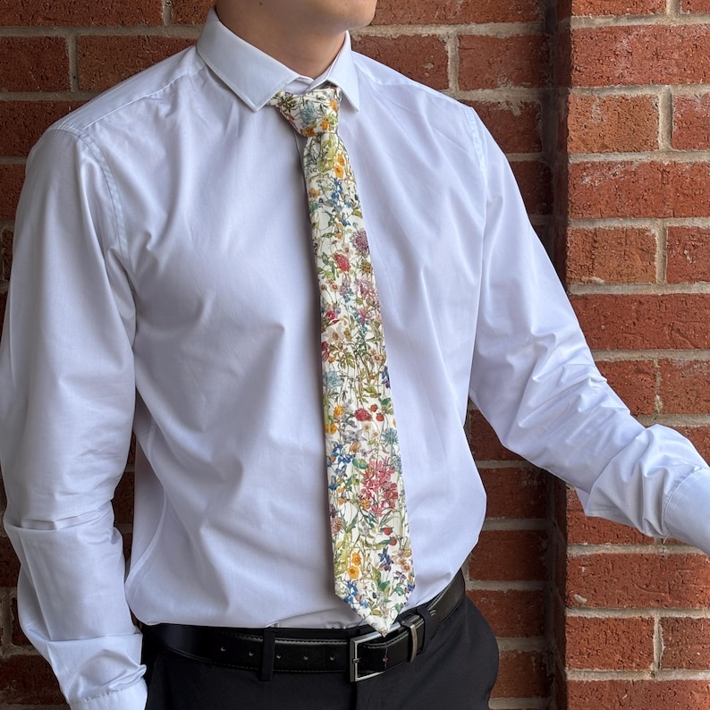 May include: A white dress shirt with a floral patterned tie. The tie features a variety of colorful flowers, including red, yellow, and blue, against a white background. The shirt is buttoned up and the person is wearing a black belt.