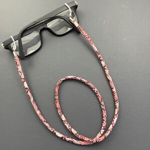 May include: A pink and white floral patterned eyeglass chain with silver metal clasps. The chain is looped around a pair of black eyeglasses.