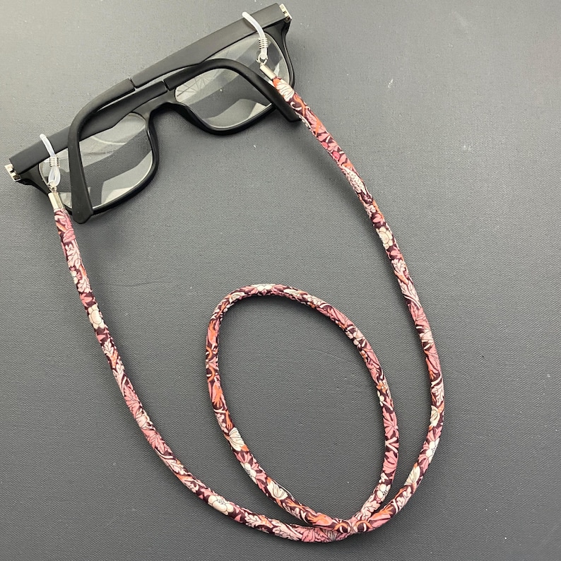 May include: A pink and white floral patterned eyeglass chain with silver metal clasps. The chain is looped around a pair of black eyeglasses.