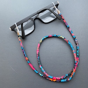 May include: Black eyeglasses with a floral patterned eyeglass chain. The chain is made of fabric and has pink, blue, and green flowers on a black background.