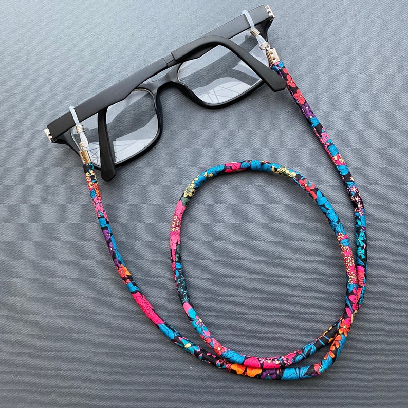 May include: Black eyeglasses with a floral patterned eyeglass chain. The chain is made of fabric and has pink, blue, and green flowers on a black background.