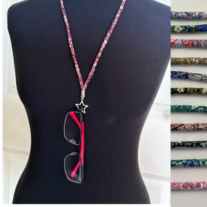Puede incluir: Un soporte para gafas con estampado floral con un dije de estrella plateada, que sostiene un par de gafas con montura roja. El soporte se muestra en un maniquí negro. Se muestran muestras de tela adicionales a la derecha.
