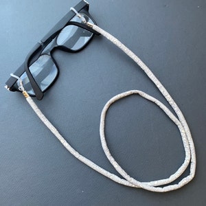 May include: A white eyeglass chain with a floral pattern. The chain is made of fabric and has metal clasps to attach to glasses.
