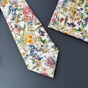May include: A white tie and pocket square set with a vibrant floral pattern. The design features a variety of colorful flowers, including pink, yellow, blue, and purple, set against a white background. The tie is angled, and the pocket square is square.