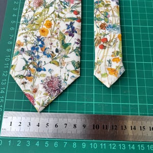 May include: Two floral patterned neckties. The ties feature a white background with a colorful botanical design of flowers and berries in shades of yellow, blue, purple, and red. The ties are placed on a green cutting mat with a ruler.