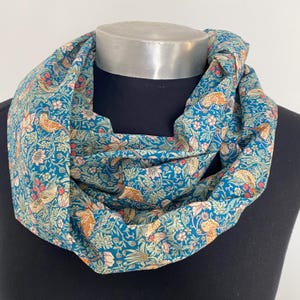 May include: A teal blue infinity scarf with a repeating pattern of stylized birds and flowers. The pattern is in shades of pink, green, and brown.
