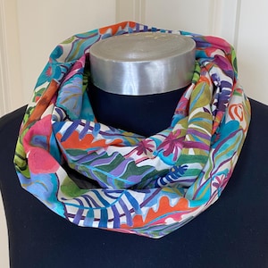 May include: A colorful infinity scarf with a white background and a tropical print featuring green, blue, purple, and orange leaves and flowers.
