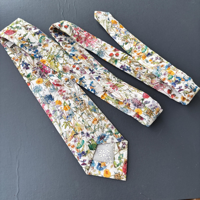May include: A set of three floral ties. The ties feature a white background with a colorful pattern of flowers in shades of pink, yellow, blue, and purple. The ties are made of a soft fabric and are suitable for formal or casual wear.
