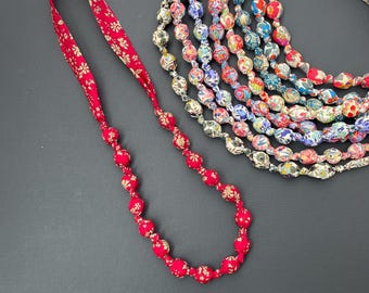 May include: A red fabric necklace with a floral pattern. The necklace is made of a series of round beads. The necklace is on a dark gray surface.