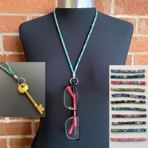 Puede incluir: Un cordón de tela azul verdoso y blanco con un anillo plateado y un llavero. El cordón se utiliza para sujetar unas gafas y una llave. La imagen también muestra una variedad de otros patrones de tela en los que se puede hacer el cordón.