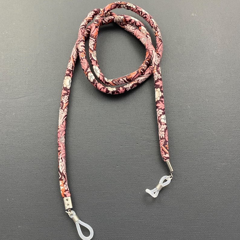 May include: A pink and white floral patterned eyeglass chain with silver metal clasps. The chain is made of fabric and is approximately 28 inches long.