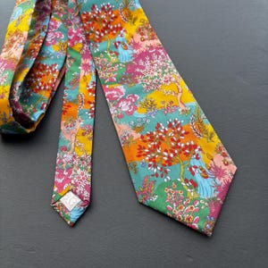 May include: A necktie featuring a vibrant, multi-coloured floral and botanical pattern. The design includes shades of yellow, teal, pink, and red, depicting stylised trees and blossoms across the fabric surface in a dense, repeating arrangement.