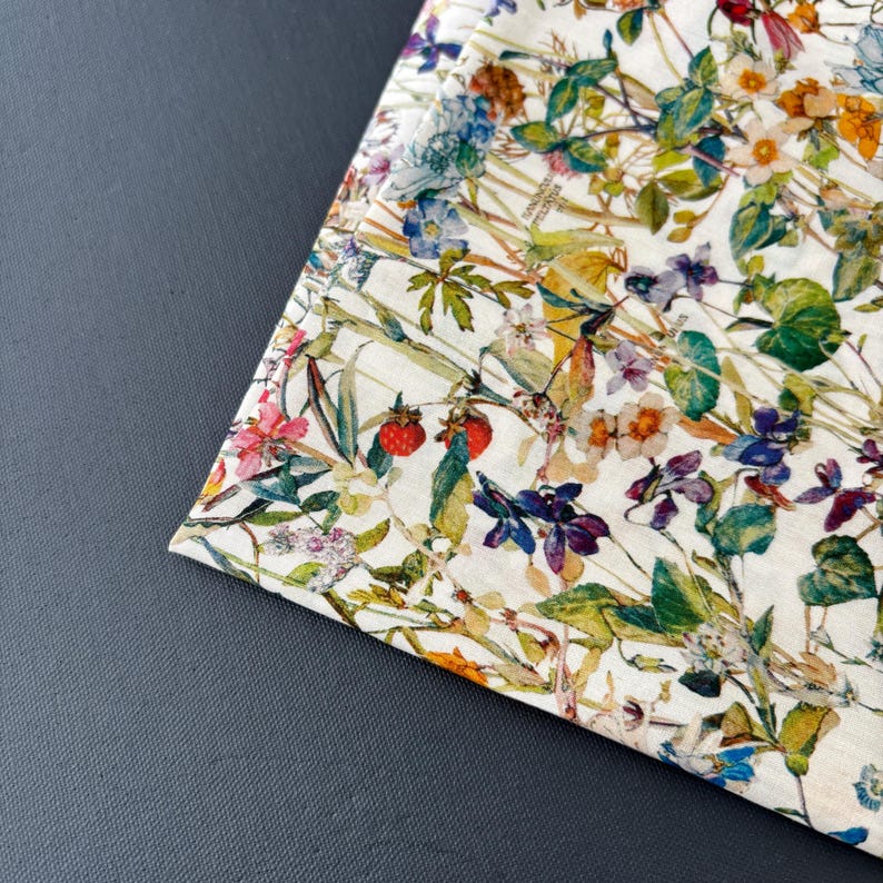 May include: A white fabric with a vibrant floral pattern featuring red strawberries, blue, purple, and orange flowers, and green leaves. The fabric is folded over, revealing a glimpse of the pattern on the underside. The design is detailed and colorful.