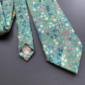 May include: A green tie with a floral pattern in shades of pink, blue, and yellow. The tie is made of a soft, cotton fabric and has a pointed tip.