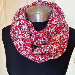 May include: A red and white floral print infinity scarf. The scarf is made of a lightweight fabric and features a pattern of small red flowers and green leaves on a white background. The scarf is displayed on a black mannequin.