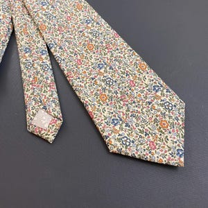 Puede incluir: Una corbata de color crema con un pequeño estampado floral en tonos azules, rosas, naranjas y verdes.