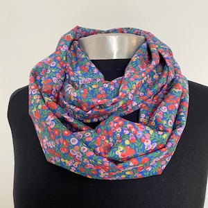 May include: A blue infinity scarf with a floral pattern. The scarf features a repeating design of small red, pink, and yellow flowers with green leaves. The scarf is displayed on a black mannequin.