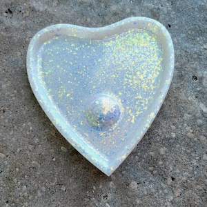 Puede incluir: Un plato blanco en forma de corazón con purpurina iridiscente incrustada en la resina. El plato tiene una ligera hendidura en el centro.