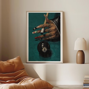 May include: A framed print showing a hand with long, decorated nails and rings, holding a pool cue above a black eight ball. The background is a teal green, similar to a pool table. The image has a close-up perspective.