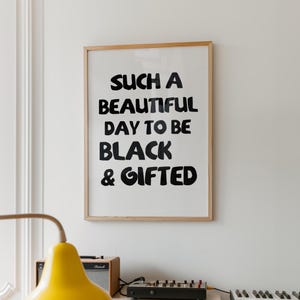 May include: A rectangular wall art print in a light wood frame featuring the black text "SUCH A BEAUTIFUL DAY TO BE BLACK & GIFTED" printed on a white background. The typography is bold, sans-serif, and hand-lettered in style, centered within the frame.