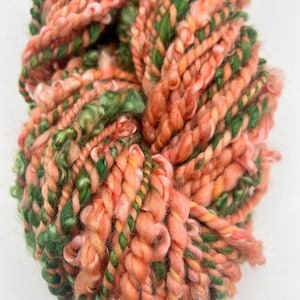 May include: A close-up of a skein of hand-spun yarn. The yarn is a mix of peach, orange, and green colors. The yarn has a thick, textured appearance, with varying thicknesses and a slightly fuzzy texture. The yarn is likely used for knitting or crocheting.