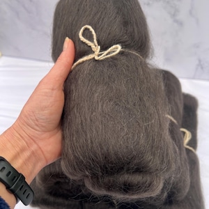 May include: Dark gray wool roving, bundled and tied with natural twine. The soft, fluffy texture of the fiber is visible, suggesting it's suitable for spinning, felting, or other fiber arts projects. The bundle is held in a hand.