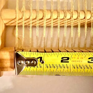 Pode incluir: Close-up de um pequeno tear de madeira com fios de fio bege. Uma fita m&eacute;trica amarela mostra que o tear tem aproximadamente 5 cm de largura. Para pequenos projetos de artesanato.