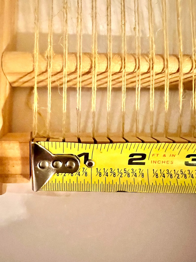 Pode incluir: Close-up de um pequeno tear de madeira com fios de fio bege. Uma fita m&eacute;trica amarela mostra que o tear tem aproximadamente 5 cm de largura. Para pequenos projetos de artesanato.