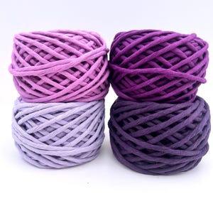 May include: Four skeins of colorful yarn in shades of purple and lavender are stacked in pairs. The yarn is tightly wound, showing the texture of the material. The color palette ranges from light lavender to deep purple.
