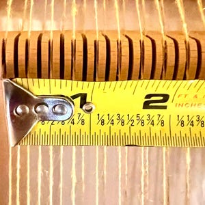 Pode incluir: Close-up de uma ferramenta de tecelagem de madeira com uma fita m&eacute;trica amarela. A fita mostra uma medida de aproximadamente 4,4 cm. A ferramenta tem ranhuras verticais e fios finos de cor clara que a atravessam.