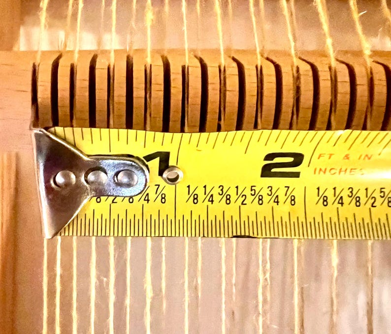 Pode incluir: Close-up de uma ferramenta de tecelagem de madeira com uma fita m&eacute;trica amarela. A fita mostra uma medida de aproximadamente 4,4 cm. A ferramenta tem ranhuras verticais e fios finos de cor clara que a atravessam.