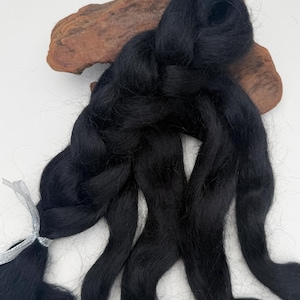 May include: A bundle of black fiber, possibly wool or yarn, with a silver ribbon tied around a portion of it. The fiber is arranged in flowing strands, with a dark brown wooden element in the background.