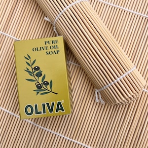 May include: A box of "OLIVA" pure olive oil soap with a green olive branch illustration. The box is gold and sits on a bamboo mat. A rolled-up bamboo mat is also in the image.