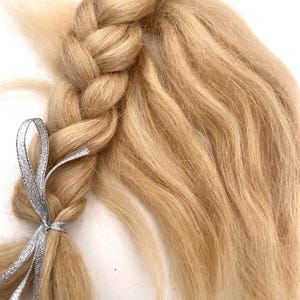 May include: A close-up of a blonde braid tied with a silver ribbon. The braid is thick and textured, with strands of hair flowing out from the side. The ribbon is thin and shiny, adding a touch of elegance to the hairstyle.
