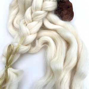 May include: Close-up of a braid of off-white wool fiber, secured with a gold ribbon. The braid is thick and textured, with loose strands flowing from the end. A piece of dark brown wood is visible at the top, providing a contrasting background.