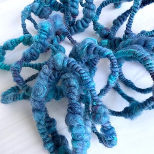 May include: Close-up of a collection of hand-spun yarn in shades of blue and teal. The yarn is thick and textured, with a slightly fuzzy appearance. The yarn is arranged in a loose, organic pattern.
