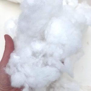 May include: Close-up of a handful of fluffy, white polyester fiberfill. The soft, cloud-like material is used for stuffing pillows, cushions, and plush toys. The fibers are tightly packed together, creating a dense, yet lightweight texture.