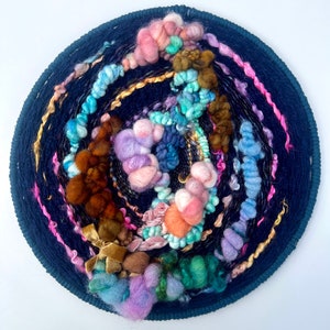 May include: A circular wall hanging with a dark blue background and a swirling pattern of colorful yarn. The yarn is arranged in a variety of textures and colors, including pink, blue, green, brown, and gold.