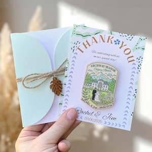 May include: A wedding thank you card and envelope set. The card features a pin with the text "Archway Manor Calgary Alberta" and the names "Rachel & Jim" with the date "09.06.2026". The envelope is tied with twine and a wooden leaf charm.