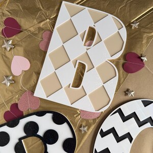 May include: A large white letter "B" with a checkered pattern and a beige background. Decorative hearts and star-shaped ornaments are scattered around the letter on a gold background. Other patterned letters are also visible.