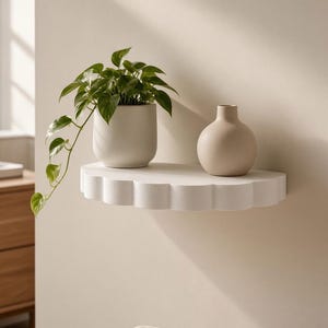 May include: A white semi-circular wall shelf with a scalloped edge. A matte white ceramic pot containing a green trailing plant sits on the left side, and a smooth, round, beige ceramic vase is positioned on the right side of the shelf.